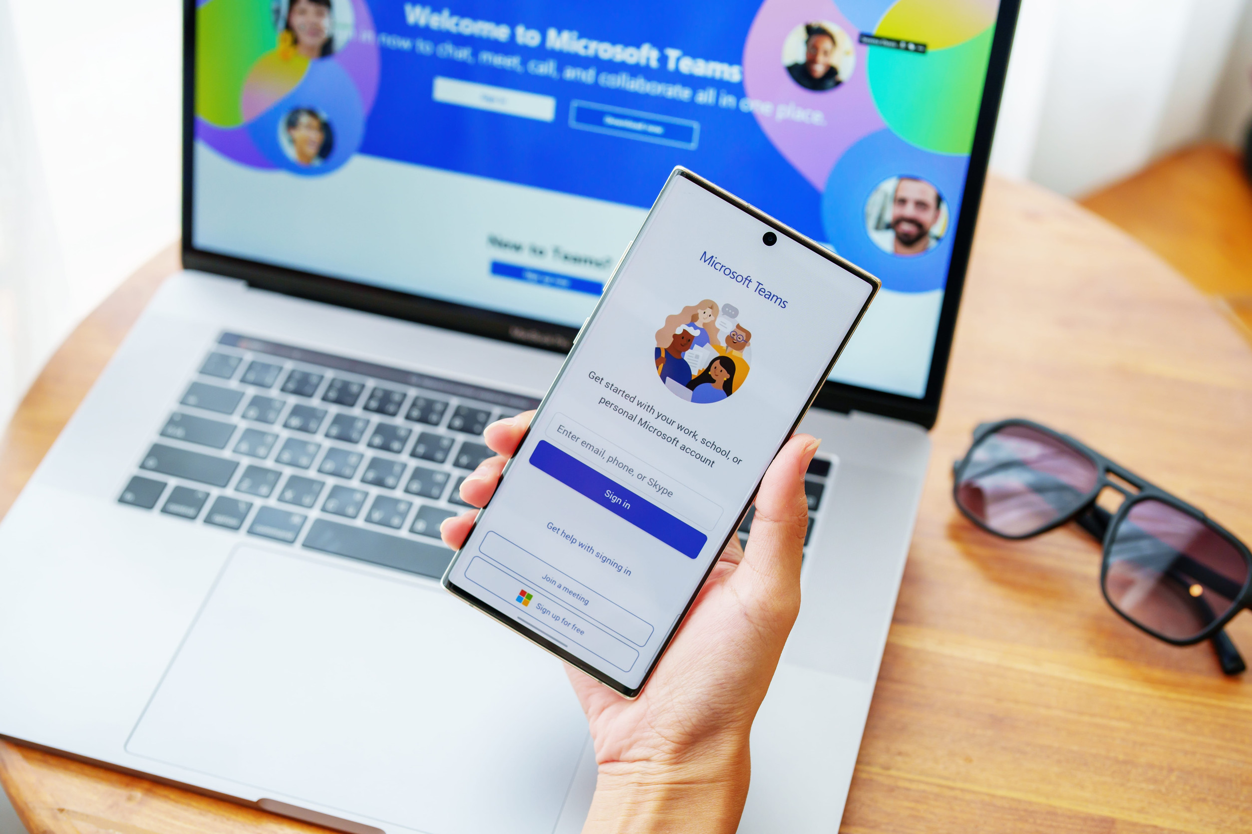 Person Holding A Smartphone Displaying The Microsoft Teams App Login Screen With A Laptop In The Background